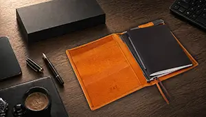 Flachgelegte Ansicht eines Schreibtischs mit einem geöffneten X47 Leder-Terminplaner in Hellbraun. Umgeben von Business-Accessoires wie einer Armbanduhr, einem iPhone, einem Füller und einer schwarzen Geschenkbox.