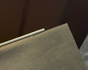 Roehrchen_am_Heft Nahaufnahme eines dünnen, glänzenden Metallröhrchens, das parallel zur oberen Kante eines grauen, strukturierten Heftcovers verläuft.