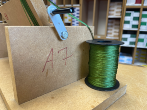 Spezial-Werkzeug Ein hölzernes Spezial-Werkzeug in einer Werkstatt, bestehend aus einer MDF-Halterung mit der Aufschrift „A7“, an der eine grüne Kordel über eine Metallführung von einer Spule abgewickelt wird.